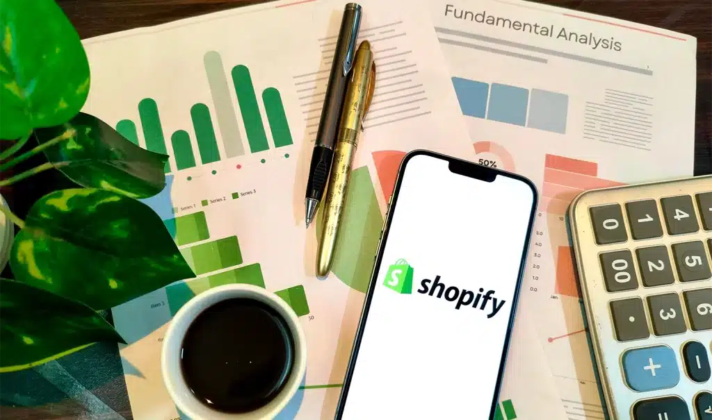 Scrivania con fogli dedicati alla pianificazione del budget, una calcolatrice e uno smartphone acceso che mostra la schermata con il logo di Shopify.